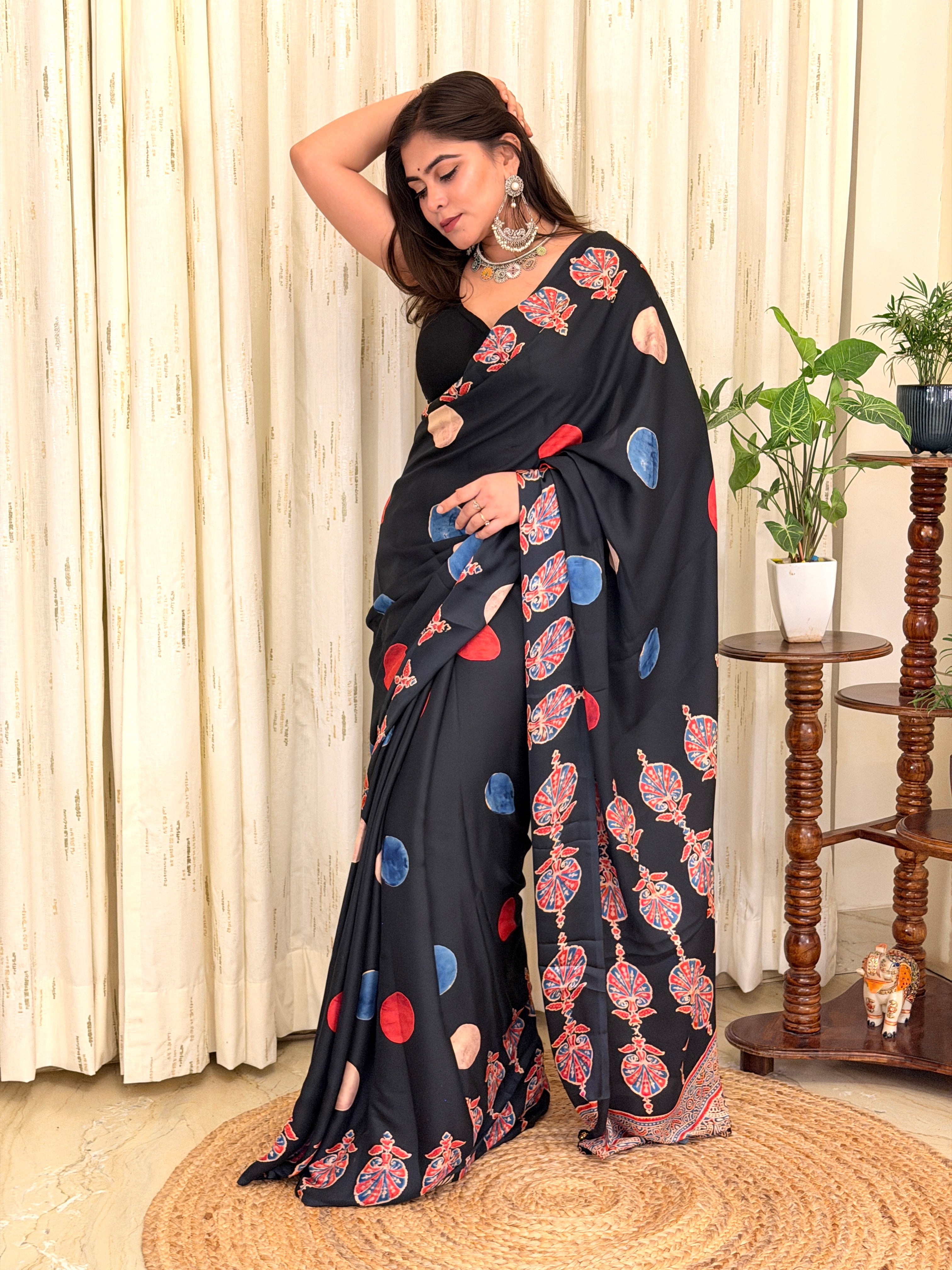 Full-length view of a woman modeling a premium black crepe saree featuring vibrant red, blue, and beige abstract polka dots and an intricate red Kalamkari motif border.