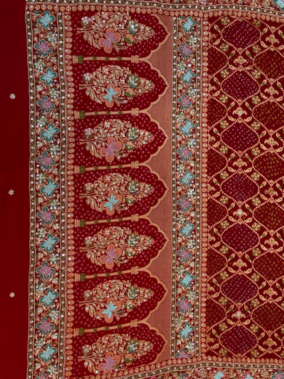 Close-up detail of a crimson red dupatta showing intricate gold zardozi, sequin, and pastel thread hand-embroidery forming large floral bouquets inside traditional architectural arches over a bandhani print.