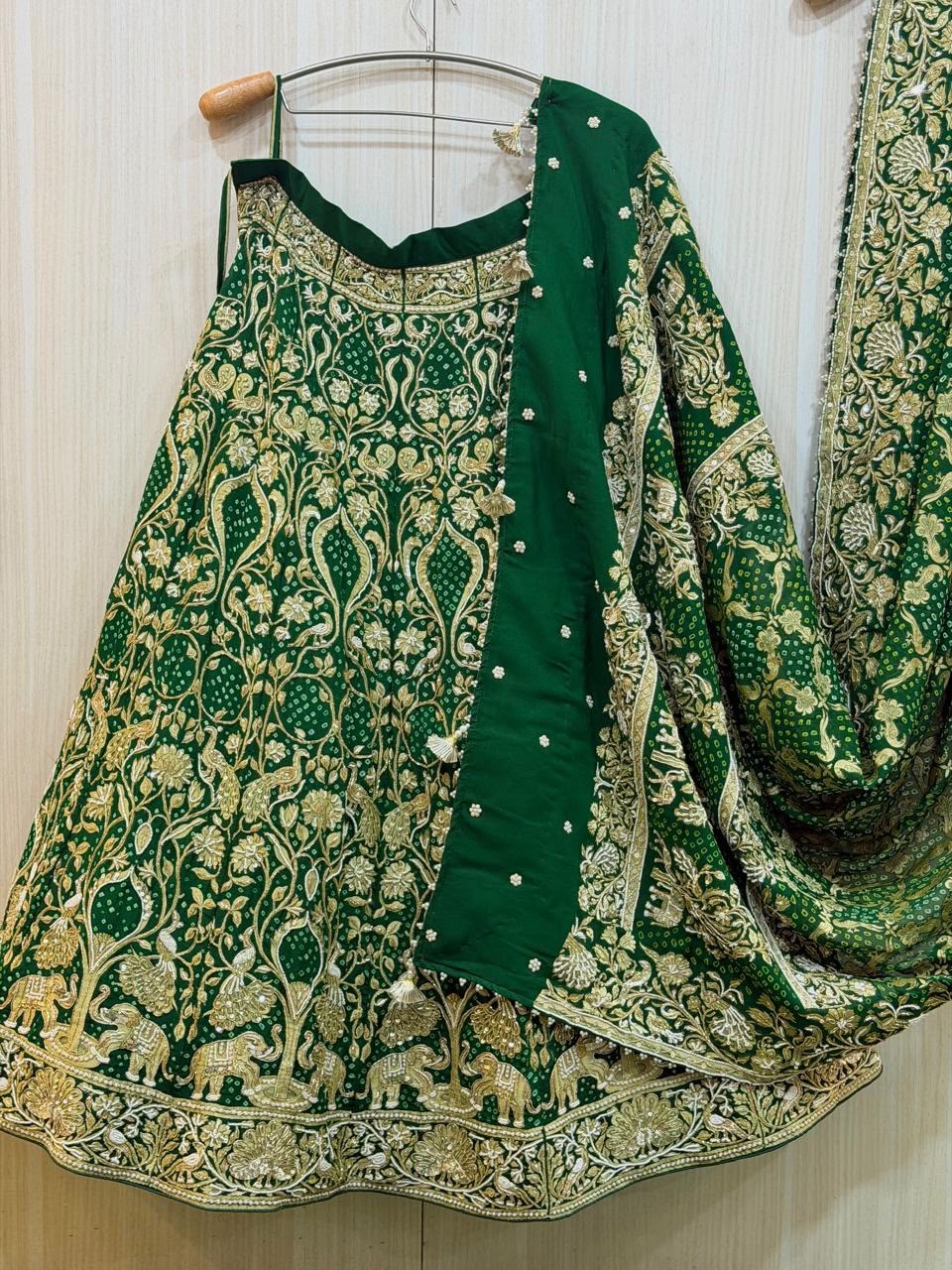 "A deep emerald green lehenga skirt and matching dupatta hanging on a wooden hanger. The outfit is heavily adorned with intricate gold embroidery, featuring a dense pattern of floral vines, peacocks, and a prominent bottom border of marching elephants. The matching dupatta has thick embroidered borders, scattered motifs, and delicate pearl tassels."